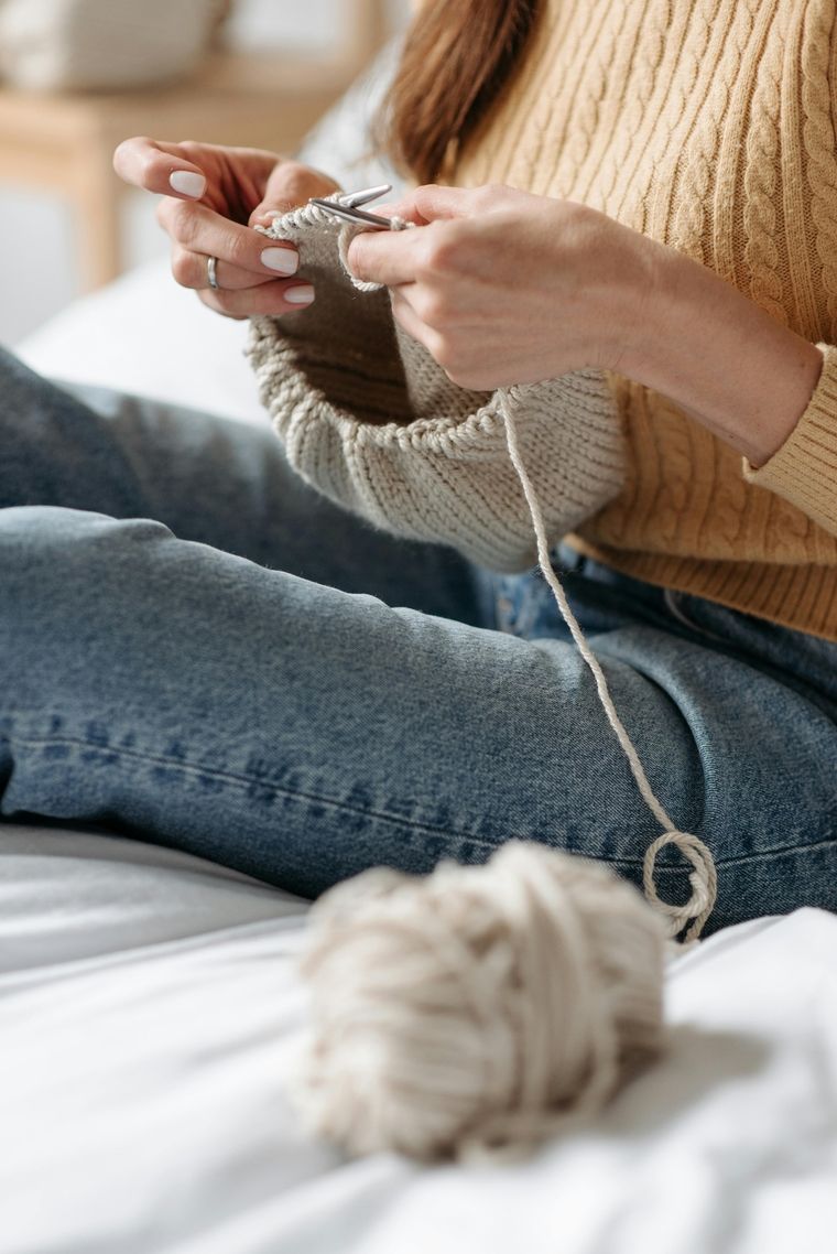Person sitzt auf einem Bett und strickt mit beiger Wolle, das Strickstück liegt auf dem Schoß